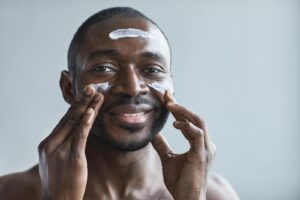 A cheerful African American man applying facial cream indoors.