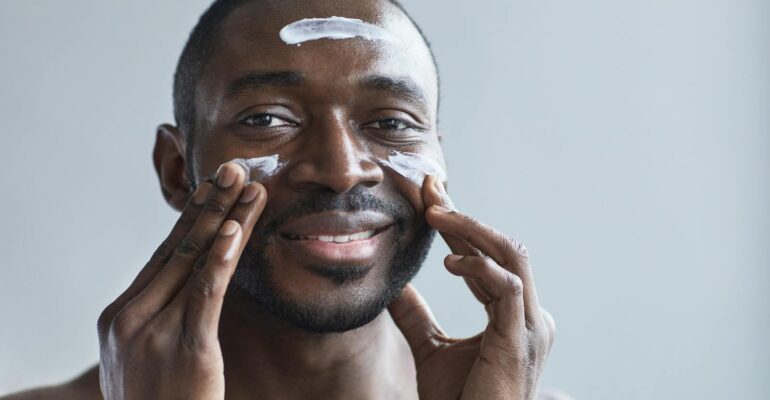 A cheerful African American man applying facial cream indoors.