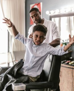 A boy with a playful expression gets a haircut from a barber. Bright indoor scene.